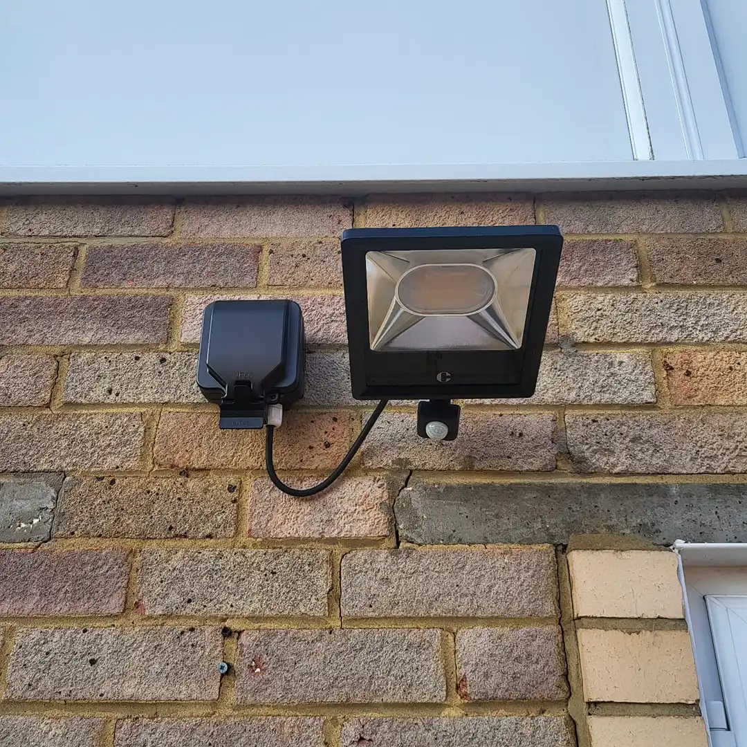 Modern black external residential light installed on a brick wall of a house with white paneling above and the corner of a door visible.